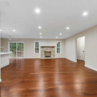Interior view of an unfurnished open-concept living room with brown hardwood floors and beige walls. Centered on the far wall is a stone fireplace with a wood mantel, centered by two tall windows. To the left, sliding glass doors open to the outside, with a ceiling fan above and the edge of a white kitchen island and gooseneck faucet are visible. Recessed lights span the ceiling, and on the right a doorway leads to a hall beside a white interior door and wall thermostat.