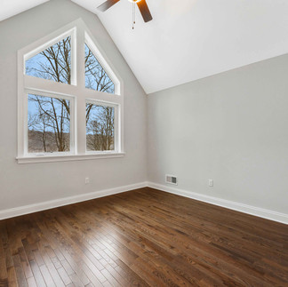 Unfurnished bedroom with a vaulted ceiling, dark hardwood floors, and a ceiling fan, with a large gable-style window with views of leafless trees outside.
