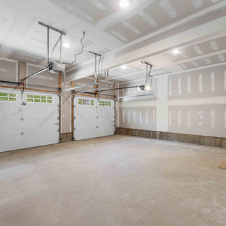 Large unfinished two-car garage with drywall, concrete floors, two overhead garage doors with window panels, ceiling-mounted openers, and a small wooden staircase leading into the home.
