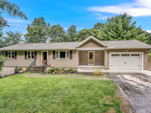 Exterior view of a single-story house in Brodheadsville, PA with beige siding, dark maroon shutters, and a garage with a white door. The entrance has a set of stairs leading to a front porch with two light fixtures on either side of the door. The yard is green with landscaping along the front, and there are trees in the background.