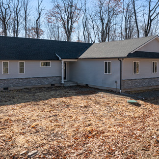 Exterior angled view of a single-story ranch-style house in Canadensis, PA with light gray siding, a dark shingled roof, and a stone veneer along the lower exterior. The house has several rectangular windows and a recessed covered entryway. The house is on a wooded lot with bare trees and a neighboring house in the background and a yard covered in dry leaves and grass.