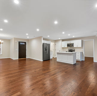 Interior view of an unfurnished open-concept living area and kitchen. The space has brown hardwood floors and light beige walls with recessed lights across the ceiling. A chandelier hangs over the dining nook by a single window, and a dark front door is to the left. The kitchen has white shaker cabinets, a center island with a sink, and stainless steel appliances, including a refrigerator, range, and microwave, with a pantry door nearby. A ceiling fan is on the far right.