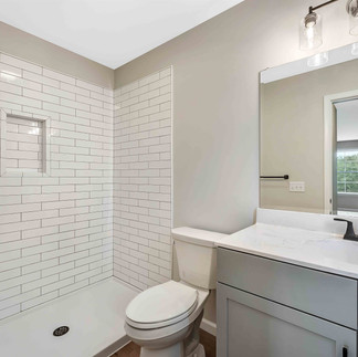 Finished bathroom with a white subway-tile shower and black fixtures, a toilet, and a gray vanity with a white countertop and black faucet. A large mirror and glass-shade light fixture hang above the sink.