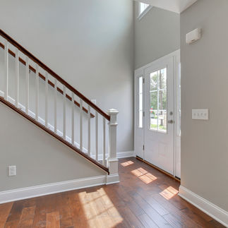 Interior view of entryway space with grey walls, white trim, dark brown wood flooring, white door an doorway with long windows on each side of the door. Stairway in front of door leading upstairs with white railings, and wood banisters.