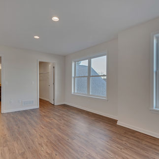Large unfurnished upstairs bedroom space with wood-look flooring, two large windows providing natural light and neighborhood views, recessed ceiling lighting, and open doorways leading into an en-suite bathroom with a vanity and a walk-in closet with built-in shelving.