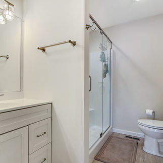 Finished bathroom with a white vanity, with a single sink, large mirror, and overhead lighting. The bathroom has a glass-enclosed walk-in shower, a toilet with a beige floor mat, and a tall white storage cabinet with glass-paneled doors.