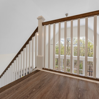 Interior view from a loft area overlooking a two-story living space with large windows, white balusters with a wooden handrail.