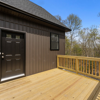 Close-up view of a wooden entry deck attached to a modern home with vertical dark brown siding. The deck has a natural wood railing and floorboards, leading to a black exterior door. A wall-mounted light fixture and electrical outlet are located beside the door. 