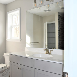 Finished bathroom with a gray vanity with a white integrated countertop and oval sink, brushed-nickel faucet, and a wide frameless mirror above the vanity topped by a multi-light fixture with clear glass shades. A window and towel bar sit on the left wall, and the entrance to a dark gray tiled shower is visible to the right. The window is providing natural light that is leaving bright rays on the wall.