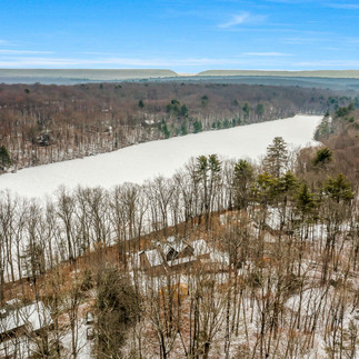 Exterior aerial view of a frozen lake bordered by snow-dusted forest, with a few homes within the trees and distant mountain ridgelines.