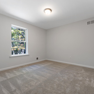 Unfurnished bedroom with light gray walls and beige carpet. The room has a window that overlooks a wooded area, providing natural light. A white door is visible on the right side, and there is an air vent on the wall near the floor. 