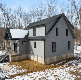 Exterior side view of a two-story gray house in East Stroudsburg, PA with a black roof, set on a wooded lot. The house has multiple windows, a covered front entry with stairs leading up, and a concrete foundation partially exposed above ground. Snow covers the yard and surrounding trees, with patches of bare ground visible near the house.
