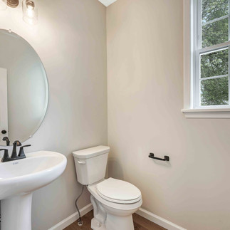 Small half-bathroom with beige walls, a white pedestal sink, and a round mirror with a black faucet. A toilet sits beside a large window, with black hardware and light wood flooring.