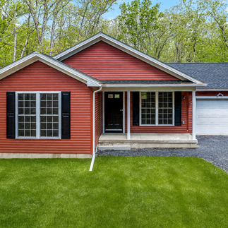 Front exterior view of a red single-story house in Bushkill, PA with black shutters and a covered porch, with a white one-car garage and gravel driveway, surrounded by tall trees and a green lawn.