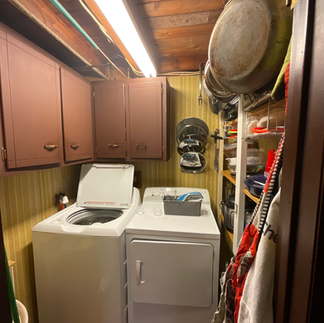 A small laundry room with a white washing machine and dryer. There is a basket placed on top of the dryer. Above the washer and dryer, there are brown cabinets and a shelf with kitchen items, including pots and pans. Various kitchen tools and cleaning items are hanging from the wall. The room has a wooden floor and is lit by a fluorescent light.