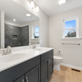 Finished bathroom with a double vanity, marble countertops, and gray cabinets. A large mirror spans the width of the vanity. The bathroom has a white toilet, towel bar, and a shower with gray stone tiles. Two windows allow natural light.