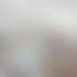 Unfurnished open-concept kitchen and living area with a central island with light-colored cabinets and a grey speckled countertop. There are pendant lights hanging above the island, and large windows provide natural light and views of a deck and trees outside. The unfurnished living area in the background has a stone fireplace and large windows, with beige walls and light wood-like flooring throughout,