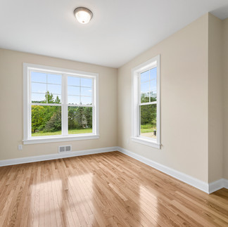 Interior view of an unfurnished room with beige walls, white trim, a white ceiling with a ceiling light, and warm brown wood-look flooring. There is a double window on the back wall and a single window on the rightside wall, bith providing natural light. An air vent and electrical outlet are seen on the back wall below the window.