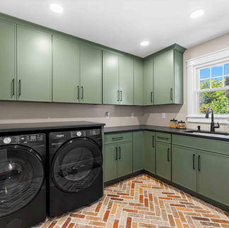 Interior laundry room with sage-green cabinets and black handles, black washer and dryer, and a black countertop with an undermount sink below a white-trimmed window. The space has herringbone flooring and recessed ceiling lights.