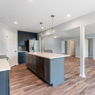 Kitchen with dark blue cabinets, stainless steel appliances, and a large center island with white quartz countertop and built-in sink. Pendant lights hang above the island, while wood-look vinyl plank flooring extends into the open-concept living area. The space has light gray walls and recessed lighting with multiple doorways leading to other rooms.