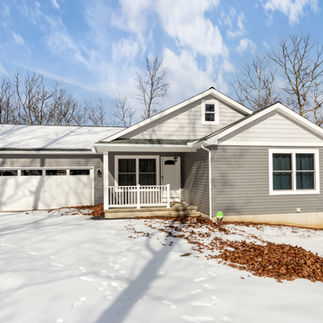 Exterior view of house with gray vinyl siding, white trim, and a covered front porch, in a snowy wooded landscape. The house has a two-car garage with windows, and black exterior lighting fixtures.