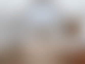 Open concept kitchen and living area with high vaulted ceilings, large windows, light hardwood floors, gray walls with white trim, a spacious kitchen island with wood cabinets, a white marble-style countertop, stainless steel appliances, and a sink under a window. Sliding glass doors lead to a small outdoor deck.