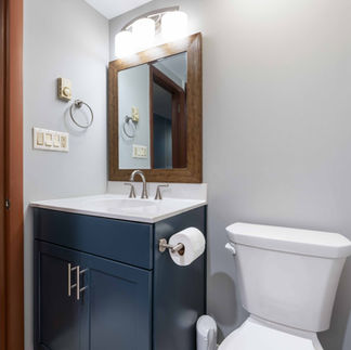 A bathroom with a dark blue vanity and a white sink. The vanity has two handles and a silver faucet. A white toilet sits next to the vanity, with a toilet paper roll holder mounted on the side of the vanity. Above the sink is a rectangular wooden-framed mirror, lit by a three-bulb light fixture. The walls are painted light gray and the floor is a light wooden color. A towel ring is attached to the wall next to the light switch and electrical outlets.
