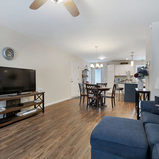 Furnished open-concept living and dining area with wood-style flooring, with a navy sectional sofa, a flat-screen TV on a media console, and a round wooden dining table with four chairs. The space leads into a kitchen with white cabinets, an island, pendant lighting, and stainless steel appliances.