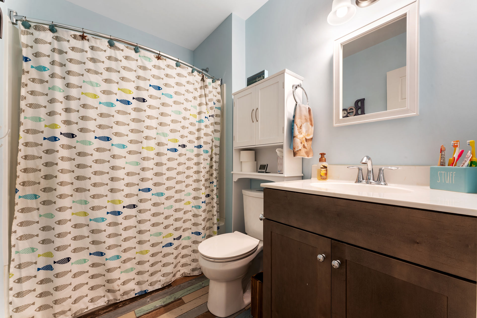 Finished bathroom with pale blue painted walls, wooden sink cabinets, square mirror above sink, white cabinets with shelf above the toilet. Full bath shower combo with closed shower curtain. 