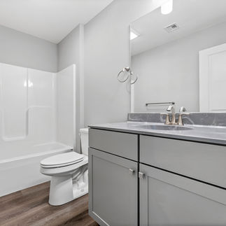 Finished bathroom with a vanity with gray cabinetry, a marbled countertop, a large wall mirror, white bathtub and shower combo with a built-in surround, a standard toilet, and brushed nickel fixtures throughout. The flooring is wood-style vinyl, and the walls are painted in a light gray.
