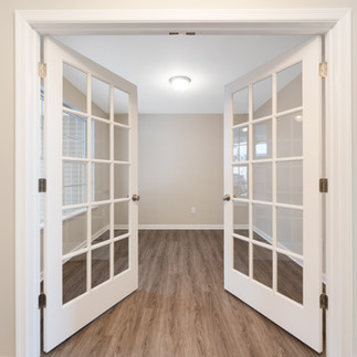 View looking into an interior unfurnished room through white french doors. The room has a window providing natural light. The walls are painted beige and there is brown wood-look laminate flooring.