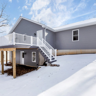 Rear exterior view of a single-story house with gray vinyl siding and a raised wooden deck with white railings and a staircase leading to a snow-covered backyard. The house has a walk-out basement with a door and window below the deck.