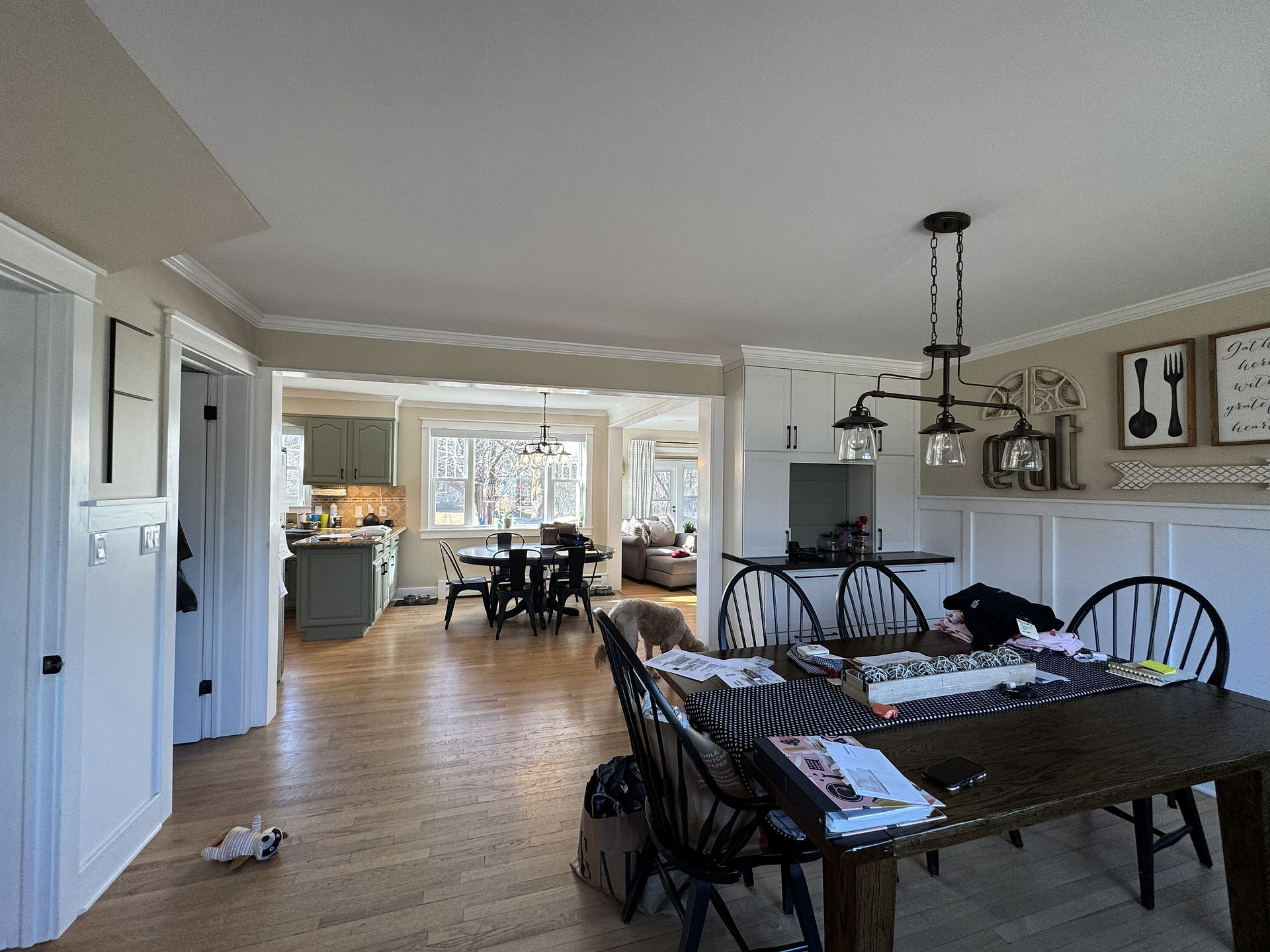 Interior view of an open-concept kitchen and dining area with hardwood floors. A dark rectangular table with papers and a centerpiece is in the foreground under a glass-shade chandelier, with a round dining table by windows in the background, gray kitchen cabinets to the left, and a dog standing near the center walkway.
