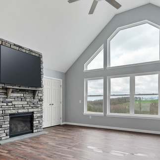 Unfurnished open living room with vaulted ceiling and large windows showing a view of green fields. The room has a ceiling fan, stone fireplace with a mounted flat-screen TV, gray walls, dark wood flooring, and a white double door closet.