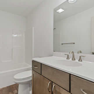 Finished bathroom. There is a white bathtub and shower combo on the left side with a chrome showerhead. There is a white vanity with a double sink, and light wood cabinets. Above the vanity is a large mirror, and a towel bar is visible to the side. The floor is a dark hardwood flooring, and the walls are painted white.