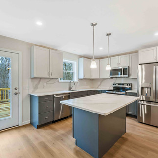 Finished kitchen with a large island, white upper and dark blue lower cabinets, quartz-style countertops, pendant lights, stainless steel refrigerator and range, and a glass door leading to a wood deck.