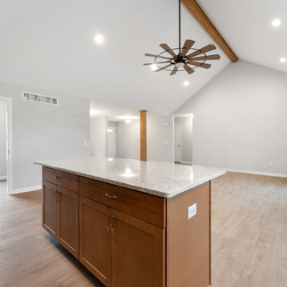 Unfurnished, open-concept living area with high vaulted ceilings, wooden beams, and recessed lighting. The room has a kitchen island with a granite countertop and wooden cabinetry. A stone fireplace is visible on the right side of the room. The walls are painted light gray, and the floor is light wood laminate. A large window in the background provides natural light.