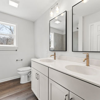 Finished bathroom with a double vanity with two rectangular mirrors with black frames above it. The vanity has a light-colored countertop with dual sinks, and gold faucets. The floor is covered with light wood laminate. A white toilet is beside the vanity, and there is a small window with a view of bare trees outside. A towel bar is mounted on the wall next to the window, and there is a shower area with tiles to the left.