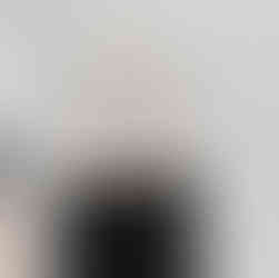 Bathroom vanity with a black cabinet and a beige countertop. The vanity has a circular sink with a gold faucet. Above the sink, there is a square mirror with a light-colored frame. The wall-mounted towel holder beside the sink is also gold. The room has white walls, and the kitchen area can be seen in the background with a stainless steel refrigerator and black cabinets. The flooring is light wood.