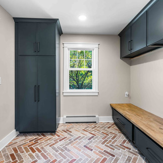 Small interior mudroom space with a herringbone floor, a tall navy storage cabinet, and a built-in wood bench with matching navy drawers beneath wall cabinets. A white-trimmed window provides views of trees.