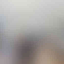 Open-concept dining and kitchen area with wood-style flooring, with a round wooden dining table with four chairs and a chandelier. The kitchen has white cabinets, stainless steel appliances, an island with pendant lighting, and a built-in microwave. There is a shelving unit with various items and a narrow console table with a floral arrangement against the right wall. A glass-panel door in the background provides access outside.