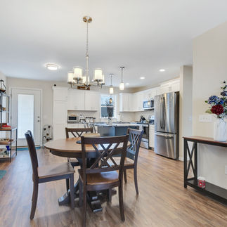 Open-concept dining and kitchen area with wood-style flooring, with a round wooden dining table with four chairs and a chandelier. The kitchen has white cabinets, stainless steel appliances, an island with pendant lighting, and a built-in microwave. There is a shelving unit with various items and a narrow console table with a floral arrangement against the right wall. A glass-panel door in the background provides access outside.