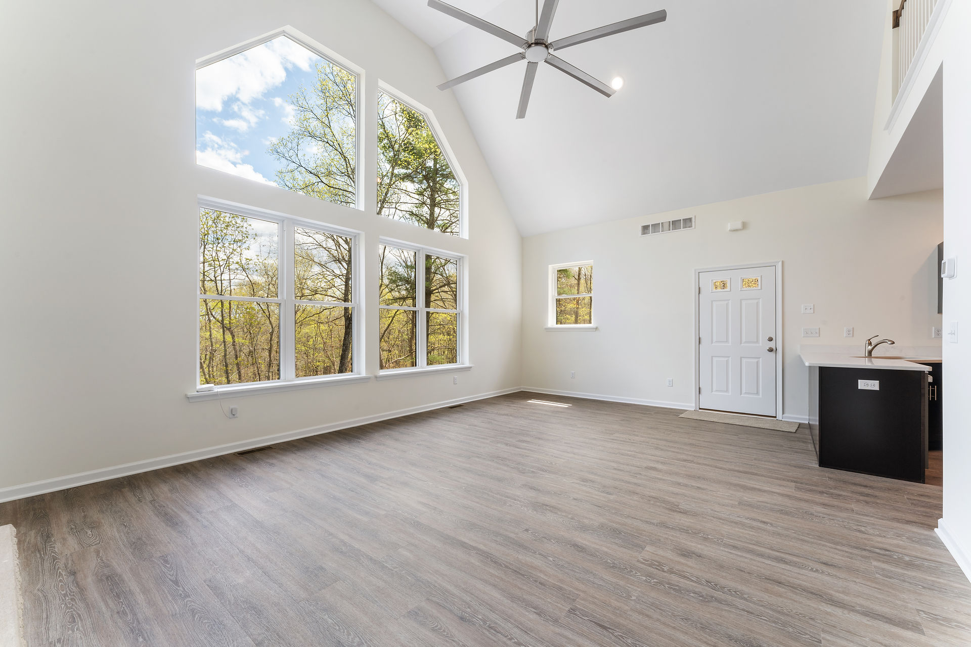 Open living room with vaulted ceilings and a large feature wall of geometric windows. The room has light-colored walls, wide-plank wood-look flooring, a ceiling fan, and recessed lighting. The entrance door and a kitchen area with a small peninsula are visible to the right. 