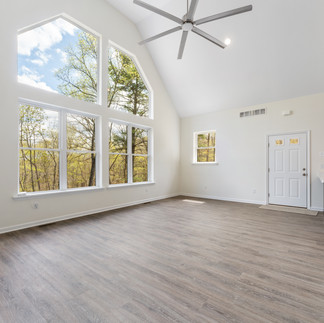 Open living room with vaulted ceilings and a large feature wall of geometric windows. The room has light-colored walls, wide-plank wood-look flooring, a ceiling fan, and recessed lighting. The entrance door and a kitchen area with a small peninsula are visible to the right. 
