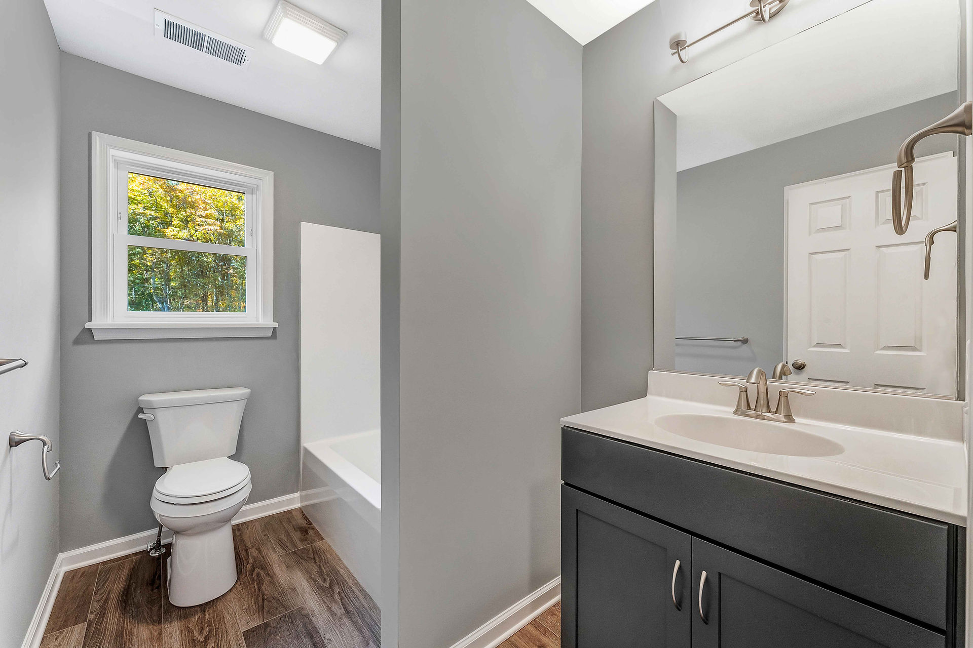 Interior view of a bathroom with gray walls, a white toilet, and a small shower stall. A vanity with a white countertop sink is installed, with a mirror above the sink and a window offering natural light.