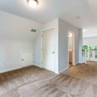 Carpeted loft area with sloped ceilings, light gray walls, and a railing overlooking large windows, with a closet door and an open doorway to a bathroom vanity.