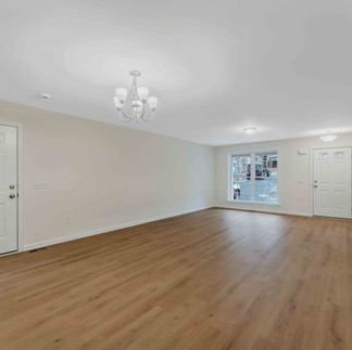 Unfurnished open-concept living and dining room with light wood floors, beige walls, a central chandelier, a large front window, and a white entry door with small glass panes.