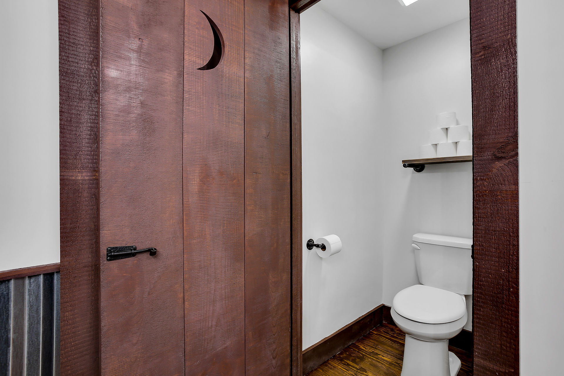 Toilet in its own seperate room in bathroom, shelf above toilet. White walls, dark drown trim, and wooden floors. Brown wooden door with crescent moon carved into door. 