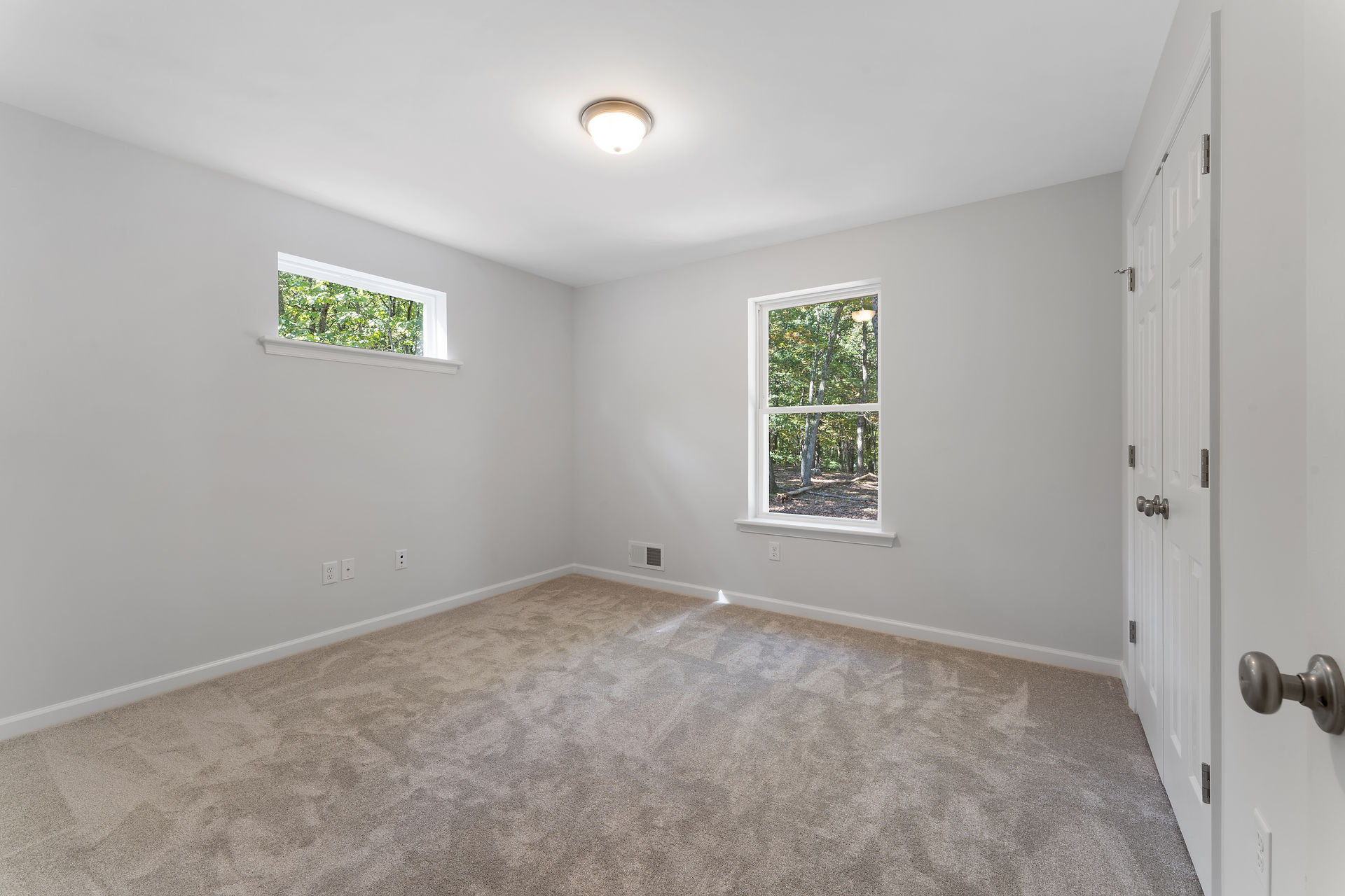 Unfurnished bedroom with light gray walls and beige carpet. The room has two windows, one with a view of greenery outside, and a door leading to a closet. There is a vent on the floor, and the ceiling is has a round light fixture.