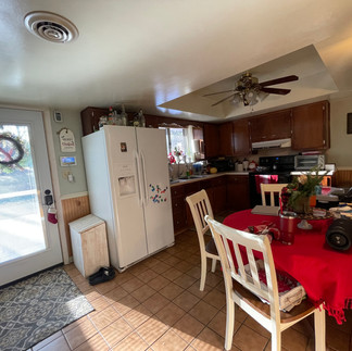 Finished kitchen with a wooden table with white chairs, covered by a red table cloth with decorative Christmas items on top, including a plant, a cup, and a speaker. On the left side, there is a white door with a wreath hanging on it, leading outside. A white refrigerator is against the wall, with small colorful magnets on it. The kitchen cabinets are brown, and the countertop has various kitchen items. Natural light is coming in through the window.